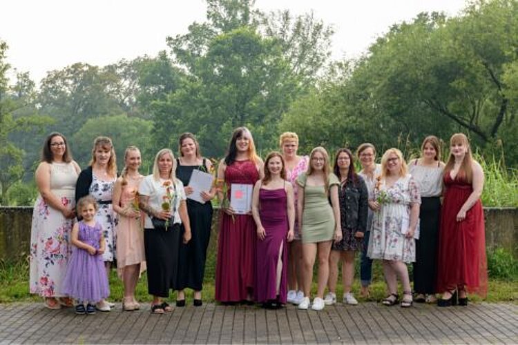 Mit Donnerschlag ins Berufsleben Eine Gruppe von 14 Schülerinnen in festlichen Kleidern und Röcken steht vor eine halbhohen Mauer, hinter der Bäume wachsen. Die jungen Frauen halten teilweise Zeugnisse und Blumensträuße in der Hand. Links steht in vorderer Reihe ein kleines Mädchen in einem lila Kleid, das offenbar die Tochter einer Schülerin ist.