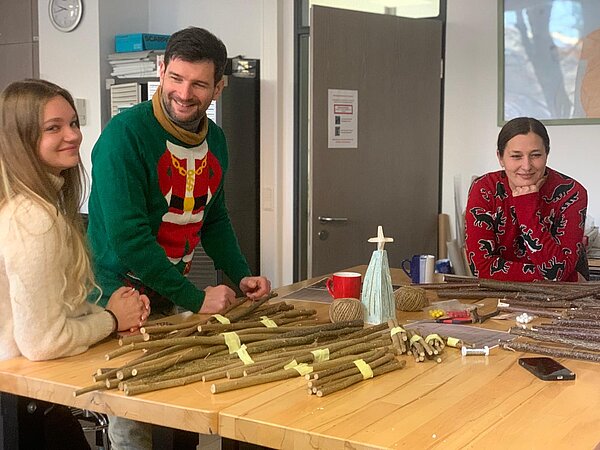 Drei Personen in weihnachtlichen Pullis stehen an einer Werkbank und lachen, vor ihnen liegen viele Holzstäbe und Bastelmaterial für dekorative Weihnachtsbäume.