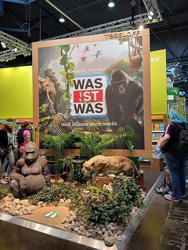 Die Leipziger Buchmesse – ein Erfahrungsbericht Der Aussteller "Was ist was" hat seinen Stand mit exotischen Tieren, Pflanzen und Steinen gestaltet. Dahinter eine große Wand mit dem Schriftzug der Marke und den gleichen Tieren