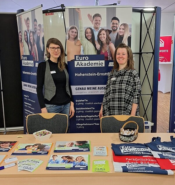 Zwei weibliche Mitarbeitende der Euro Akademie stehen lächelnd an ihrem Messestand.