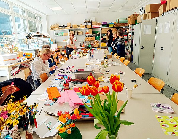 Ein heller Werkraum voller Bastelmaterialien, Tulpen und Deko-Ideen. Mehrere Frauen sitzen oder stehen an einem langen Tisch und arbeiten kreativ mit Papier, Stoff und Naturmaterialien.
