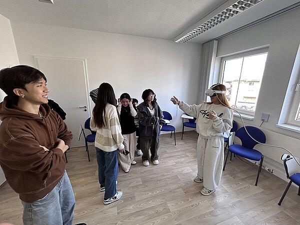 Eine Gruppe junger Auszubildender sitzt ungezwungen in einem Raum mit Holzboden und blauen Stühlen an der Wand. Eine Person rechts trägt eine VR-Brille und gestikuliert, während die anderen zusehen und lächeln. Die Atmosphäre wirkt entspannt und informell, was auf ein lockeres Treffen oder eine VR-Demonstration hindeuten könnte.