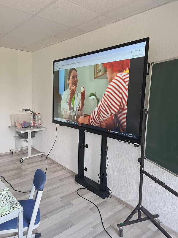 Auszeichnung „Clever in Sonne und Schatten“ 2024 Der Bildschirm hinter dem Lehrerpult zeigt einen Ausschnitt aus einem Lehrfilm, in dem gerade eine Ärztin mit einem Clown spricht.