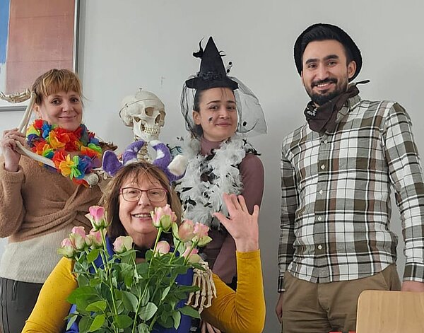Vier Personen in festlichen Verkleidungen posieren in einem Raum. Eine Frau mit Blumenkranz und einem Skelettkopf in der Hand, eine andere mit Hexenhut und Federboa, ein Mann mit einem Halstuch sowie eine ältere Dame mit violetten Plüsch-Ohren und einem Strauß rosa Rosen. Alle lachen fröhlich in die Kamera.