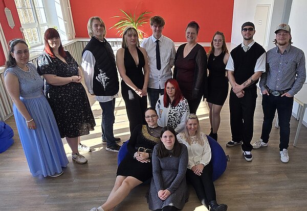 Mottowoche der Abschlussklassen Sozialassistent*in und Erzieher*in Gruppenfoto in einem hellen Raum mit roter Wand. Die Teilnehmenden tragen elegante Outfits – von Kleidern bis Hemd mit Krawatte. Einige sitzen auf Sitzsäcken vorne, die Stimmung ist festlich.