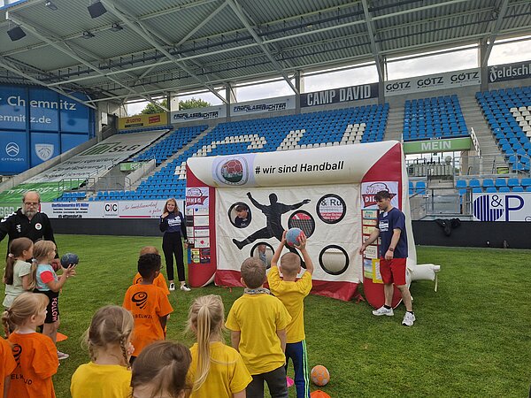 Kinder werfen bei einer Mitmachstation im Stadion Bälle auf eine Handball-Zielwand, begleitet von Mitarbeitenden der Euro Akademie Chemnitz.
