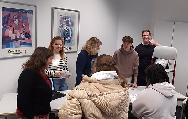 Eine Gruppe Schüler steht um einen Tisch in einem Klassenzimmer, ein Schüler zeigt Daumen hoch.