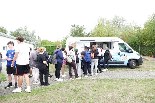 Eine Gruppe von Menschen, die teilweise Rucksäcke auf dem Rücken tragen, steht in einer Schlange vor einem blauen Food-Truck. Der Truck hat ein buntes Design mit einer Zeichnung von Speiseeis. Die Personen warten darauf, bedient zu werden.