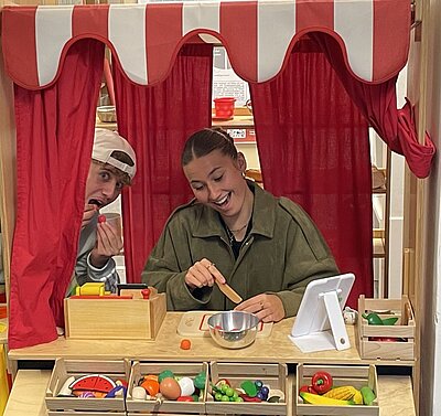 Zwei Personen an einem Spielzeug-Verkaufsstand: Die Person links mit weißer Kappe tut so, als esse sie gerade etwas, die Person rechts mit Holzlöffel lächelt über einer Metallschüssel. Auf dem Tisch und in den Fächern liegt Holzspielzeug-Lebensmittel; oben ziert ein rot-weißer Vorhang den Stand.