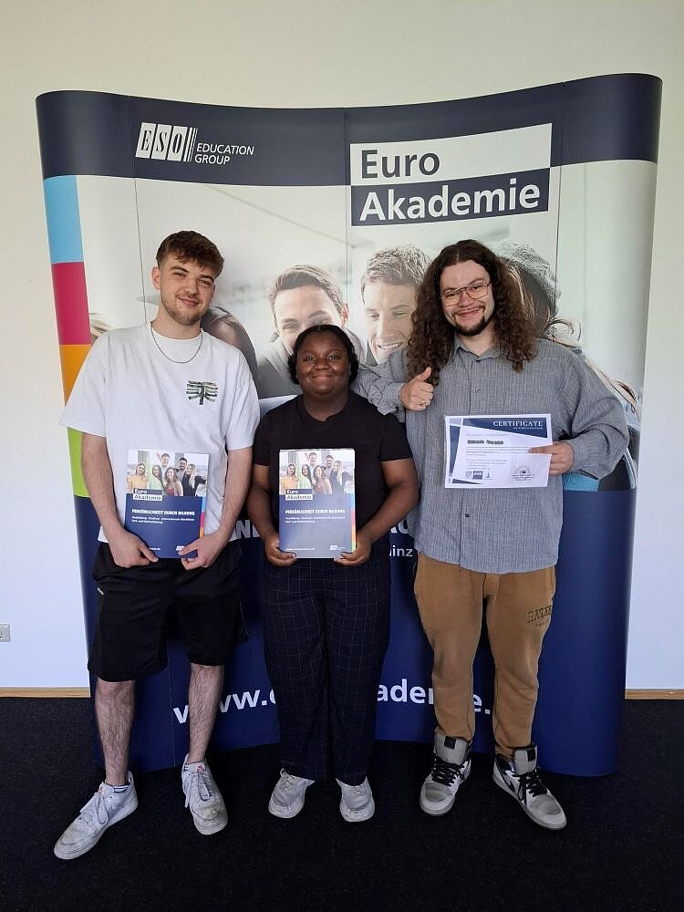 Zertifikatsübergabe Erasmus+ Drei junge Erwachsene stehen vor einem Banner der Euro Akademie Mainz und halten stolz ihre Erasmus+-Zertifikate in den Händen. Alle drei lächeln, einer zeigt den Daumen nach oben.