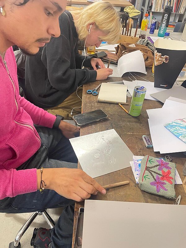Zwei Personen sitzen an einem Holztisch im Atelier: Die eine schaut auf eine bearbeitete Metallplatte, die andere zeichnet mit einem Stift auf Papier. Auf dem Tisch liegen eine Schere, ein Stift, eine Getränkedose, Papier und ein besticktes Mäppchen.