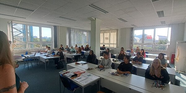 Die neuen Schülerinnen und Schüler sitzen in ihrem Klassenraum und hören ihrer Lehrerin zu.