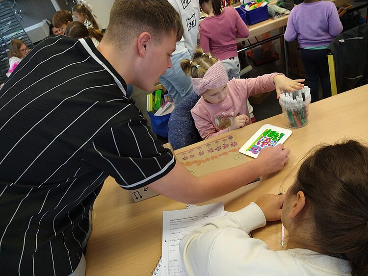 Ein junger Mann beugt sich über einen Tisch und hilft einem Mädchen beim Malen und Verzieren einer Bastelarbeit; bunte Stifte und bemalte Pappe liegen auf dem Tisch, während im Hintergrund andere Kinder basteln.