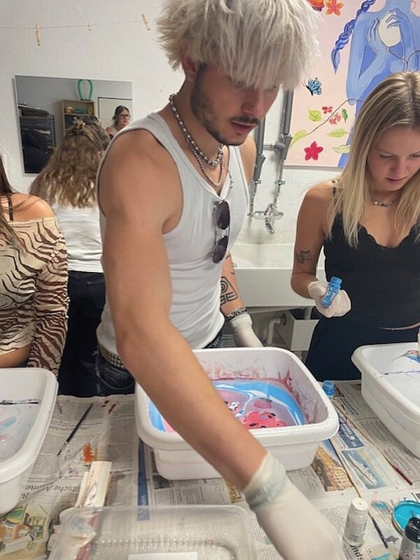 Zwei Personen arbeiten gemeinsam an einem Tisch. Eine Person mit weißem Tanktop und auffälliger Frisur greift etwas, das vor einer weißen Kunststoffwanne mit blauer und roter Flüssigkeit liegt. Die andere Person, mit blondem Haar, arbeitet an ihrer eigenen Wanne. Im Hintergrund sind weitere Materialien und ein Waschbecken zu sehen.