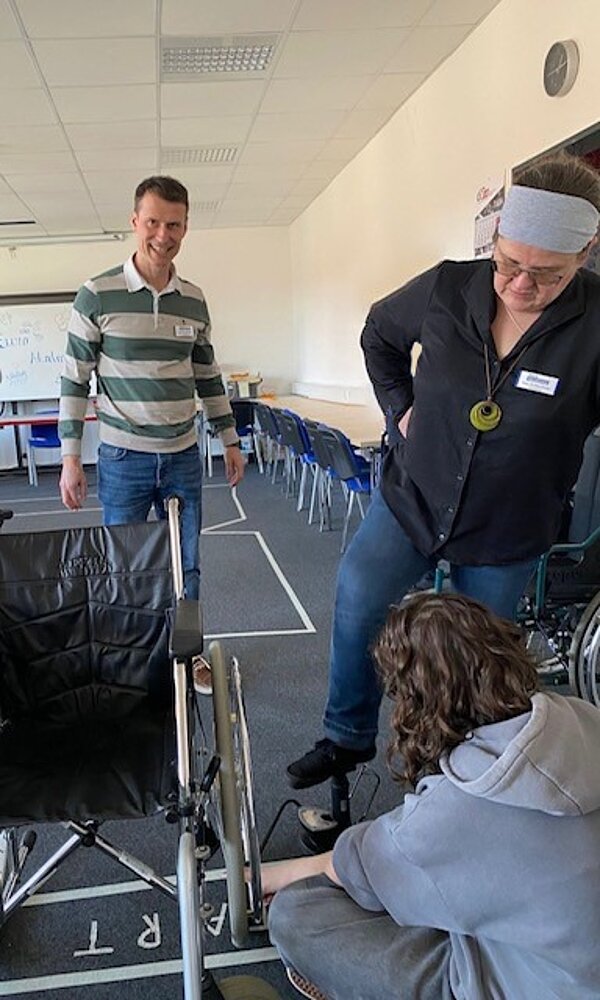 Vier Personen in einem Raum, in dem eine Übung mit einem Rollstuhl stattfindet. Eine Person sitzt auf dem Boden und hält den Schlauch einer Pumpe an den Reifen des Rollstuhls, während eine andere Personen daneben steht und mit dem Fuß die Pumpe bedient. Zwei weitere Personen begleiten die Situation. Im Hintergrund sind Stühle und Tische zu sehen.