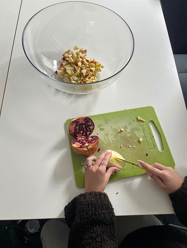 Eine Azubine schneidet auf einem grünen Schneidebrett ein Stück Apfel. Daneben liegt ein halbierter Granatapfel. Im Hintergrund steht eine große Glasschüssel mit bereits klein geschnittenen Apfelstücken auf einem weißen Tisch.