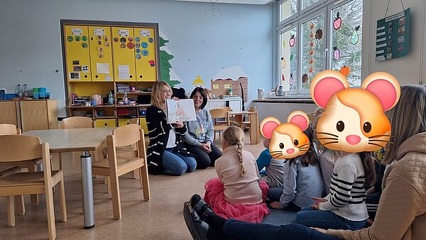 Vorlesesituation im Kita-Raum: zwei Erwachsene halten ein Bilderbuch hoch, Kinder sitzen im Halbkreis davor (Gesichter teils verdeckt).