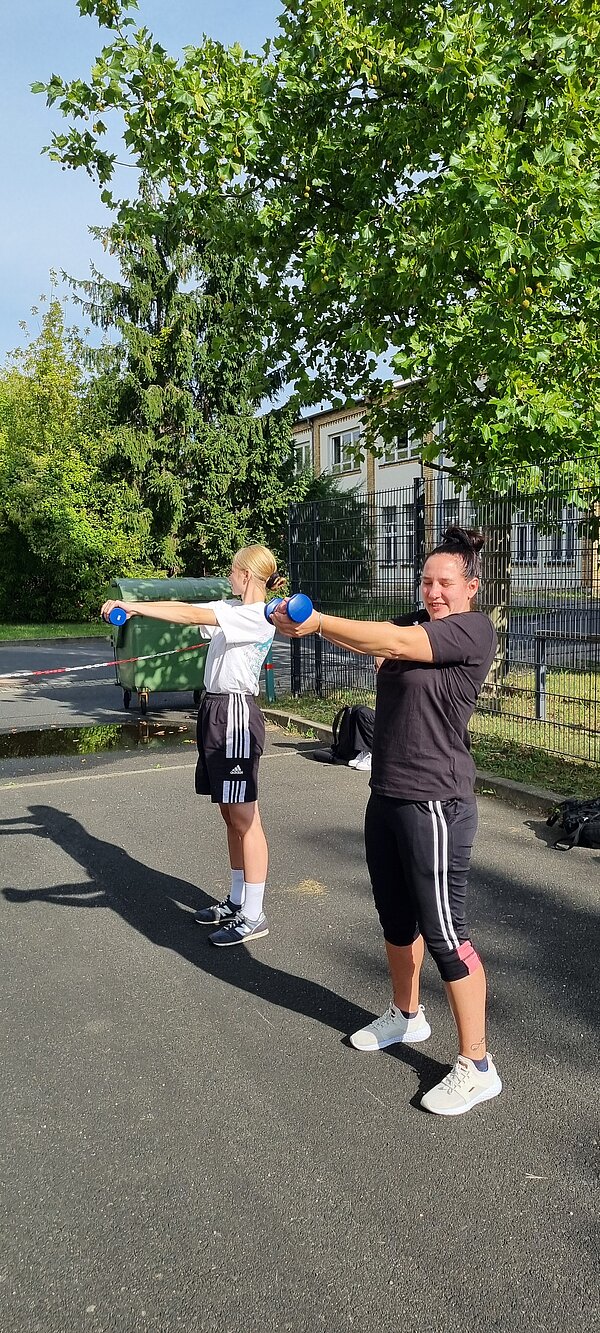 Wikinger-Sportfest: Pflegeazubis erobern die nordische Arena Zwei Frauen trainieren draußen mit blauen Hanteln. Sie stehen nebeneinander und halten die Gewichte mit ausgestreckten Armen nach vorne.