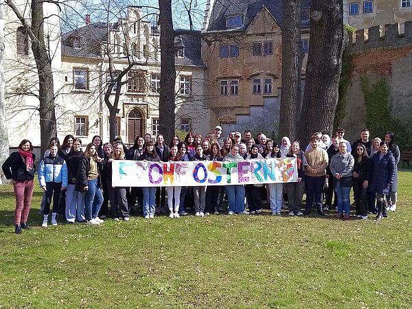 Eine Gruppe von Schülern steht auf einer Wiese vor schlossartigen Gebäuden und hält ein großes Banner mit bunten Buchstaben, auf dem Frohe Ostern steht.