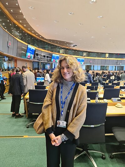 Eine junge Frau steht lächelnd in einem Konferenzraum, vermutlich in einer politischen oder institutionellen Umgebung. Sie trägt eine beige-schwarze Strickjacke, ein gestreiftes Oberteil und hat eine beige Tasche über der Schulter sowie ein Namensschild um den Hals. Im Hintergrund sieht man mehrere Personen, Tische mit Bildschirmen und eine große runde Decke mit eingelassenen Lichtern.
