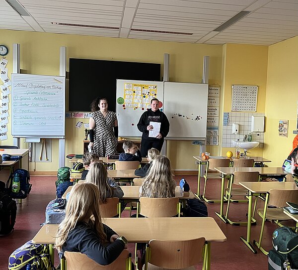 Zwei Personen stehen vorne im Klassenraum und halten Blätter in der Hand, während die Kinder an ihren Plätzen zuhören; neben der Tafel steht ein Flipchart mit Stichpunkten.
