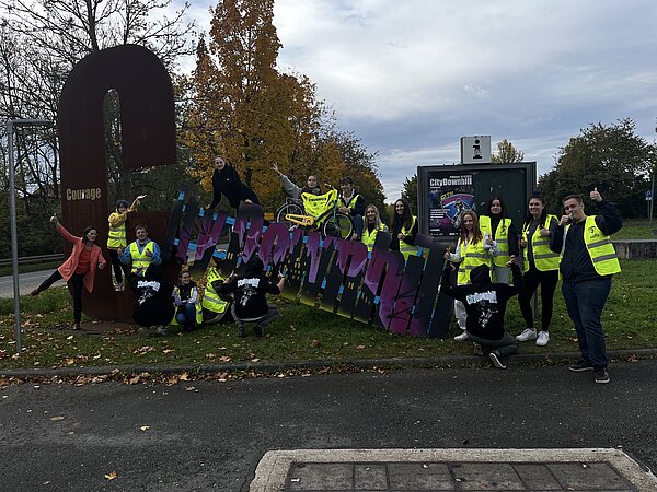 City Downhill in Pößneck Die Schülerinnen und Schüler der Euro Akademie Pößneck stehen mit gelber Warnweste auf einem Grünstreife an einer Straße. In der Mitte halten sie ein Plakat mit lila Buchstaben fest.