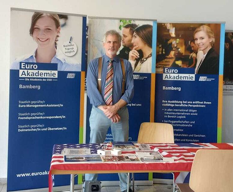 Ein Mitarbeiter der Euro Akademie Bamberg steht hinter einem Informationsstand mit Broschüren und Flyern. Im Hintergrund informieren drei Roll-up-Banner über Ausbildungsangebote in den Bereichen Management, Fremdsprachen und internationale Tätigkeiten.