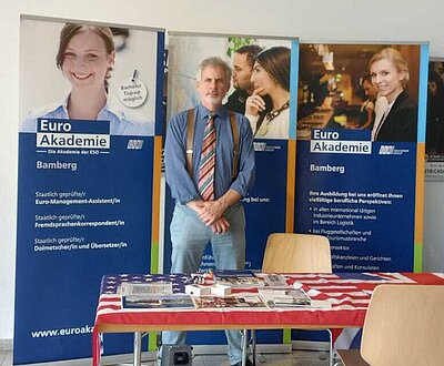 Ein Mitarbeiter der Euro Akademie Bamberg steht hinter einem Informationsstand mit Broschüren und Flyern. Im Hintergrund informieren drei Roll-up-Banner über Ausbildungsangebote in den Bereichen Management, Fremdsprachen und internationale Tätigkeiten.