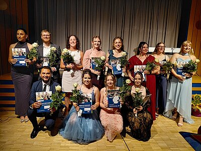 Die Absolventinnen und Absolventen posieren gemeinsam in festlicher Kleidung, mit Blumen und ihren Abschlusszeugnissen in der Hand, für ein Erinnerungsfoto.