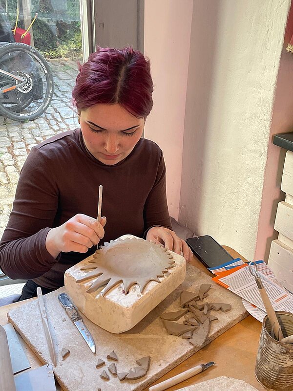 Junge Frau mit dunkelroten Haaren bearbeitet konzentriert eine Tonform in einer Gipsform. Auf dem Tisch liegen Tonreste und verschiedene Modellierwerkzeuge.