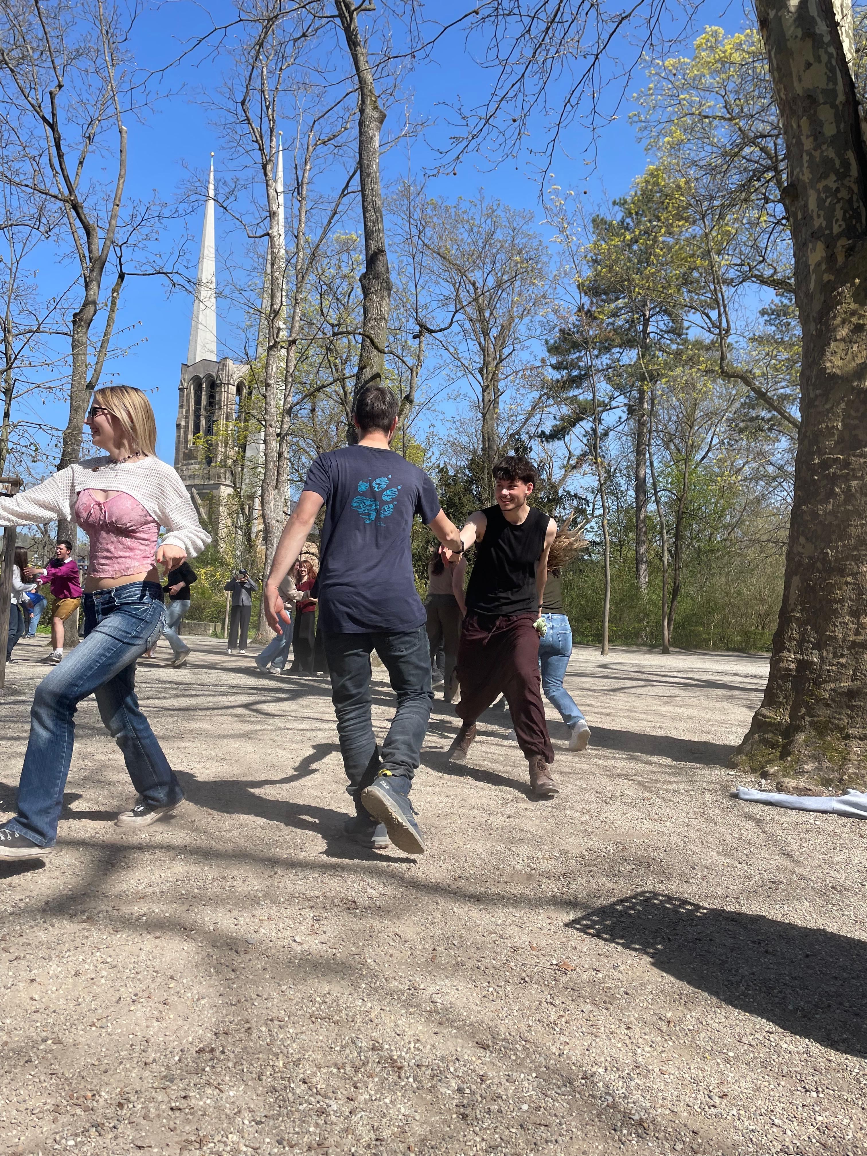 Menschen tanzen bei sonnigem Wetter im Freien Hand in Hand in einem Kreis; im Hintergrund ragen die Türme einer Kirche zwischen den noch kahlen Bäumen hervor.