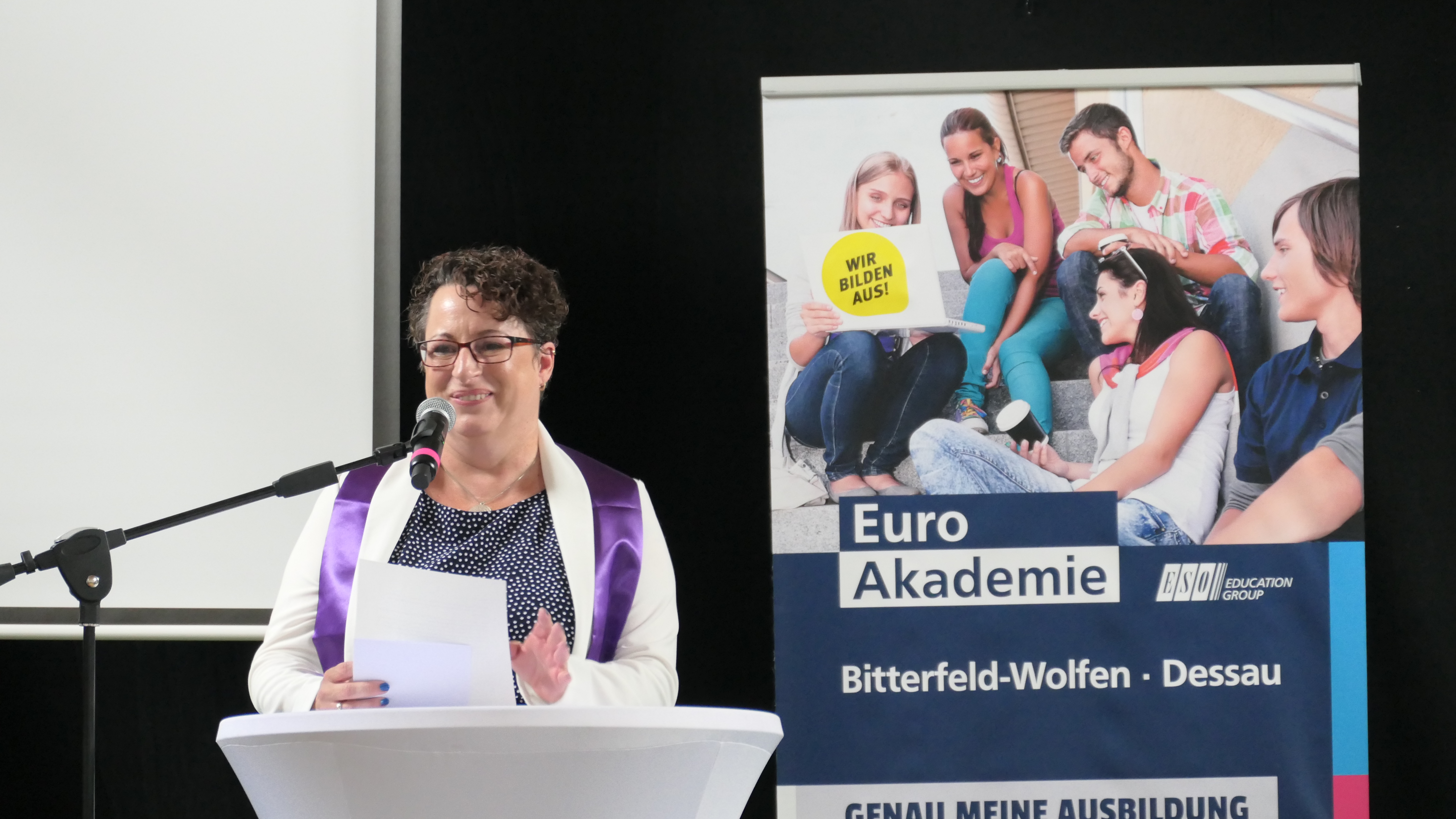 Eine Frau mit lila-weißem Oberteil steht an einem weißen Stehtisch und hält eine Rede. Im Hintergrund ist ein Werbeplakat der Euro Akademie Dessau.