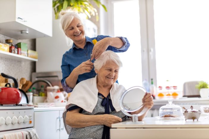 Eine lächelnde Frau schneidet die Haare einer älteren Dame, die in einer hellen Küche sitzt. Im Hintergrund sind Küchenschränke und verschiedene Haushaltsgegenstände zu sehen, was eine warme und einladende Atmosphäre schafft.