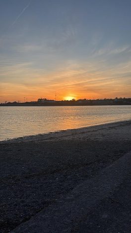 Blick vom kieseligen Strand auf die untergehende Sonne und die Skyline.