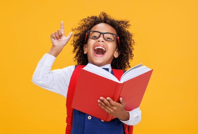 Ein kleiner Junge mit wilden, dunklen Locken steht vor einer knallgelben Wand In der linken Hand hält er ein dickes, rotes Buch, mit der rechten Hand reckt er den Zeigefinger mit gespielter Strenge nach oben. Er trägt eine dicke, schwarze Brille und hat den Mund weit geöffnet. Sein Hemd mit Weste trägt zu dem Bild des strengen Lehrers bei.