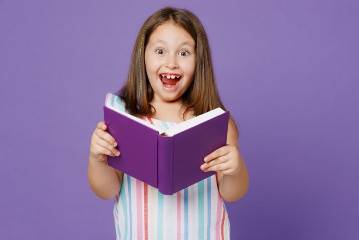 Ein kleines Mädchen in einem bunt gestreiften Kleid hält ein lila Buch in der Hand, das farblich mit dem Hintergrund harmoniert. Sie hat Augen und Mund weit aufgerissen, sodass man ihre Zahnlücken gut erkennt. In ihre glatten, braunen Haare ist eine bunte Strähne eingeflochten.