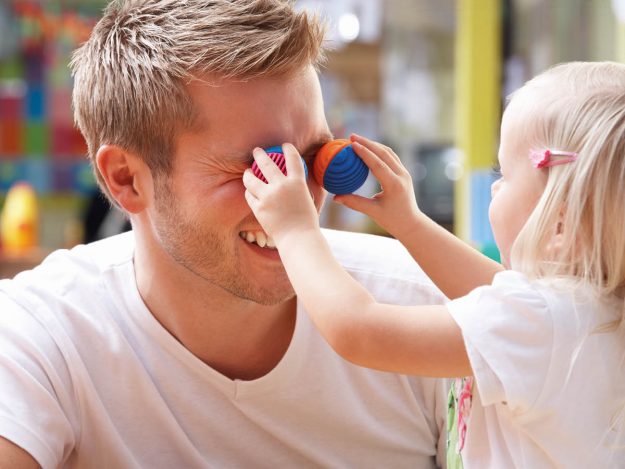 Ein kleines Mädchen hält einem Kinderpfleger zwei kleine bunte Bälle vor die Augen.
