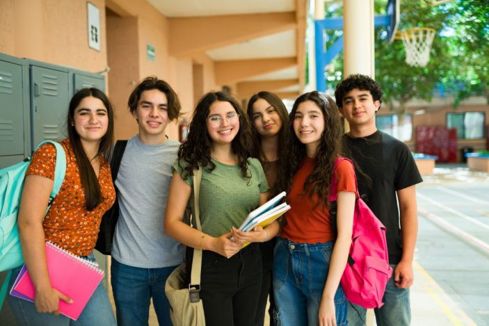Eine Gruppe von sechs lächelnden Jugendlichen steht eng beieinander in einem Schulkorridor. Sie tragen Schulmaterialien wie Bücher, Hefte und Rucksäcke und wirken entspannt und freundlich. Im Hintergrund sind Schließfächer und ein Basketballkorb zu sehen.