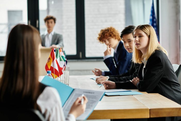 Deine Berufsaussichten mit Sprachausbildung: Diplomatie, Anwaltskanzleien und internationale Unternehmen Rechts im Bild sitzen drei junge Menschen in Anzügen an einem Holztisch; sie tragen Anzüge und schauen konzentriert nach vorne. Auf dem Tisch steht eine Reihe verschiedener Flaggen. Links im Vordergrund sitzt eine Person, die sich Notizen in einer Mappe macht. Im Hintergrund ist eine Person an einem Sprecherpult zu sehen; in der rechten hinteren Ecke hängt eine weitere Flagge.
