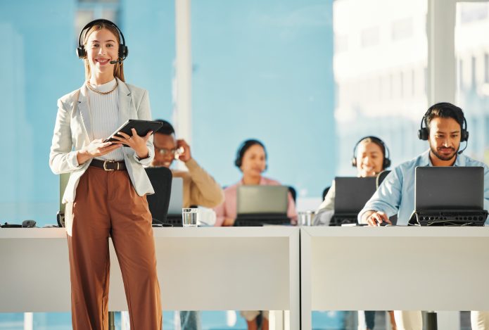 In einem hellen, luftigen Büro sitzen mehrere Personen mit Headsets an Schreibtischen mit Laptops. Vorne links steht eine lächelnde Frau mit Headset auf dem Kopf und einem Tablett in den Händen.