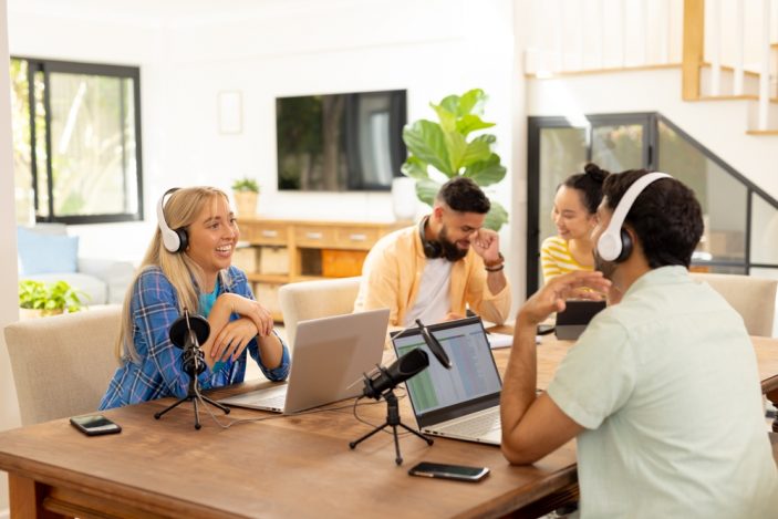 Hier sind kreative Sprachtalente gewünscht: Film, Gaming und Marketing In einem hellen Raum sitzen vier lachende junge Menschen um einen Tisch. Zwei davon tragen Kopfhörer und haben ein Mikro und einen Laptop vor sich stehen. Im Hintergrund ist ein Wohnzimmer mit Fernseher und Sofa zu sehen.