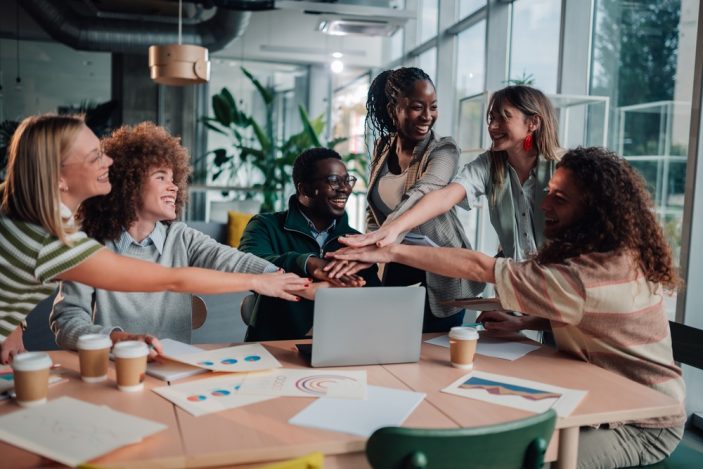 Motivation verstehen: Warum Autonomie, Kompetenz und Verbundenheit im Arbeits- und Lernalltag entscheidend sind Eine Gruppe lachender Menschen legt ihre Hände alle in der Mitte zusammen. Die Menschen sitzen und stehen um einen Tisch in einem hellen, modernen Büro. Auf dem Tisch stehen ein Laptop und Kaffeebecher, dazwischen liegen Papiere mit bunten Bildern.