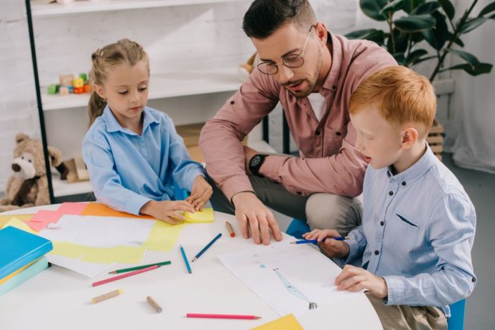 Typische Mythen über den Beruf im Faktencheck: Erzieher*in Ein Erzieher in rosa Hemd mit Brille sitzt gemeinsam mit zwei Kindern in blauen Hemden an einem runden Tisch. Auf dem Tisch liegen buntes Papier, Stifte und Bastelutensilien; im Hintergrund ein Spielzeugregal und eine große Zimmerpflanze.