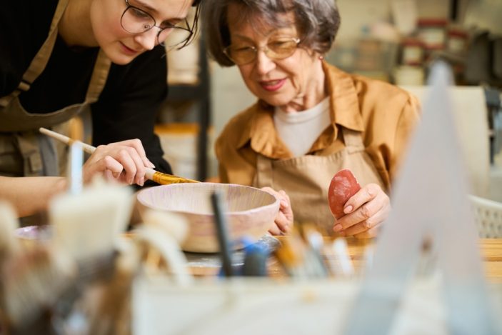 Typische Mythen über den Beruf im Faktencheck: Ergotherapie Zwei Frauen in einem Keramikstudio, die gemeinsam an einer Schüssel arbeiten. Rechts sitzt eine ältere Dame mit Brille und Schürze, die einen roten Gegenstand in der Hand hält. Links steht eine junge Ergotherapeutin, die sich herabbeugt und die Schüssel mit einem Pinsel bemalt. Im Bildvordergrund sind unscharf Pinsel und weitere Gegenstände zu sehen.